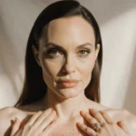 Angelina Jolie looks at camera with her hands on chest showing mastectomy scars and soft warm light highlighting empowerment