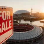 For Sale sign stands over Arrowhead Stadium with a new domed arena rising behind Kansas City skyline at sunset.