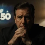 Chevy Chase looking disappointed with clasped hands dim lamp shadows a blurred SNL50 logo in background