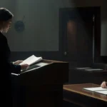 Lawyer holding sealed folder at podium with judge staring at desk in dim courtroom and partially open door