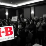 Council speaker addresses worried community members holding signs with fluorescent lights and an H‑E‑B logo in foreground