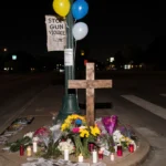 Cross stands in a small memorial with flowers, balloons, and a faded Stop Gun Violence banner in Dallas