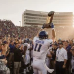 DeSoto football player holding championship trophy with cheering teammates and AT&T Stadium backdrop