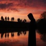 Lone figure standing with lake reflection and crimson twilight sky in background.