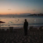 Lone figure standing by water edge on Bondi beach with sunset sky and scattered shattered glass showing quiet tragedy.