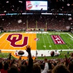 Fans cheer in stadium with split-screen fields showing Oklahoma orange and Alabama crimson football with blurred scoreboard.