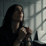 Ghislaine Maxwell looks up at her bound hands and chains in a dim prison cell with a faint petition on a table