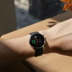 Pixel Watch 4 rests on wooden desk with soft light illuminating its bright display and autumn leaf hint in background