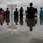 Figure stands on Bondi Beach looking toward ocean with reflection of Hanukkah crowd near a menorah