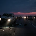 Police flashlight beams onto wrecked highway with twisted metal while emergency responders gather and sunrise light rises