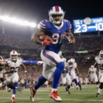 James Cook sprinting toward end zone with ball while stadium lights glow and scoreboard reads 23-20