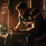 Jason Kelce sitting on a bench staring at his phone with empty water bottles and football gear in a dim Chiefs locker room.