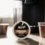 Keurig coffee machine showing a spotlighted McCafe Premium Roast pod with warm sunlight and a few surrounding K‑Cups.
