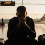 Man sits at airport gate clutching hands with scattered luggage and soft golden light casting long shadows