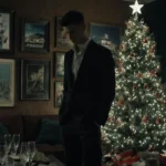 Young man staring down with empty champagne glasses amid a dim living room with Hollywood memorabilia and a Christmas tree.
