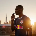 Michael Bradley coaching with a whistle and Red Bulls jersey and the New York City skyline and Red Bull Arena behind him