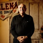 Quintanilla stands with clasped hands and a smile beside a wooden door with a faded