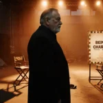 Rob Reiner standing alone with faded film poster of Being Charlie and warm orange glow behind