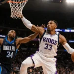 Russell Westbrook dribbles past defender with ball and left hand on rim amid Kings and Mavericks jerseys in NBA arena