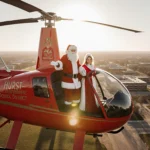 Santa Claus standing proudly inside a red Bell helicopter with Mrs. Claus beside him above sunny North Texas school grounds.