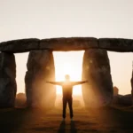 Lone figure stretches arms toward sunrise over Stonehenge stones with golden light and rising mist.