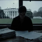 Person standing looking down at partially filled tax return with stack of financial newspapers and faint White House in backg