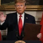 Trump stands at podium holding leather‑bound arms package with U.S. flag and Taiwanese flag in background