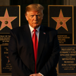 President Trump poses with arms covering face near gold‑lettered plaques on the Presidential Walk of Fame in golden hour.