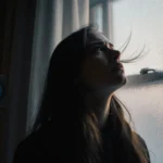 Woman staring out at frosted window with wind blowing her hair and a thermostat reading 65°F in background