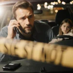 Father standing beside his car holding a phone with police tape in foreground and Christmas lights reflected on windshield.