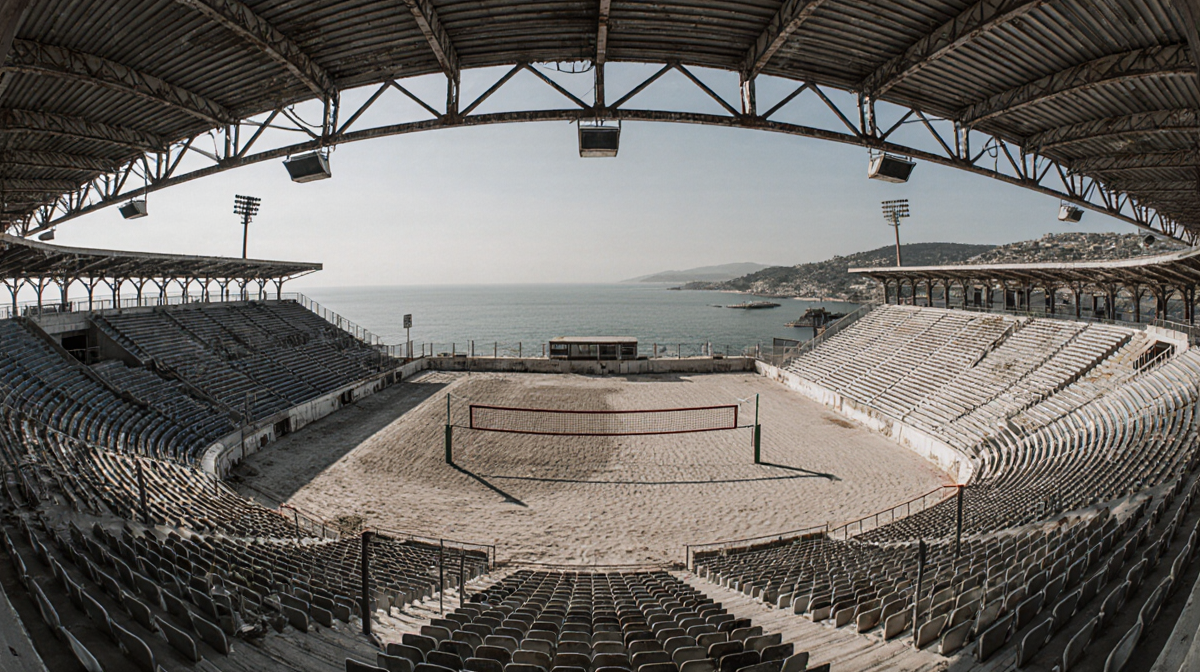 Abandoned volleyball stadium lies empty with peeling paint and rusted nets beside calm Athens coastline