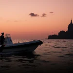 Coast Guard boat approaches Venezuelan coast with speedboat carrying two officers and presidential palace silhouette behind