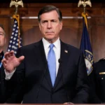 Congressman gestures toward American flag with ICE agents wearing QR code badges showing tension during immigration policy an