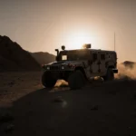 Military vehicle driving away through dusty desert terrain with plume of smoke rising and orange sunset sky