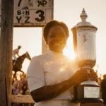Diane Crump celebrating victory with silver trophy and golden sunset light near vintage Kentucky Derby post