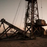 Oil derrick stands with rusted lattice and vines climbing its sides near broken wooden crane on dusty Venezuelan ground