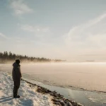 Lone figure stands at frozen Great Lakes shoreline with golden dawn light casting long shadows across snow