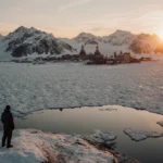 Lone figure stands at frozen lake edge with Greenland mountains glowing orange at sunset and distant mining operation visible
