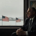 Greenlandic official sits at desk with US flag visible through window and map showing US name scribbled in red ink