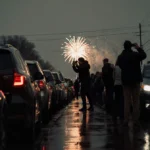 Person taking photos of gridlock cars with neon lights reflecting off wet pavement and fireworks in distant sky