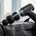 Massage gun resting on a worn gray gym bag with speed settings visible and light highlighting its portable design