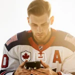 Jake Oettinger holding a worn‑out Olympic puck and gazing with Team USA gear and a American flag and maple leaf backdrop
