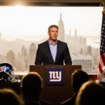 John Harbaugh stands at podium with Giants helmets and Manhattan skyline behind him