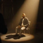 Karamo Brown shows surprise and disappointment under a spotlight with an empty chair beside him in a dim television studio.