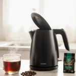 Electric kettle steams over counter with coffee cup beside it and tea bags nearby