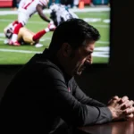 Kyle Shanahan sits with bowed head and clasped hands as blurred TV shows George Kittle injury during 49ers Eagles playoff gam