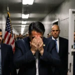 Venezuelan President Maduro walking through a Brooklyn detention center hallway with hands covering face and a U.S. flag behi