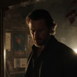 Marty Supreme stands before cracked mirror with bloodstains and film set visible behind him