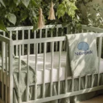 A white crib rests among sage and moss plants with soft sky blue bedding and Naturepedic logo on a basket in gentle sunlight