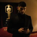 Nick Jonas stands alone in dim hotel suite with phone in hand and Golden Globes red carpet visible behind him